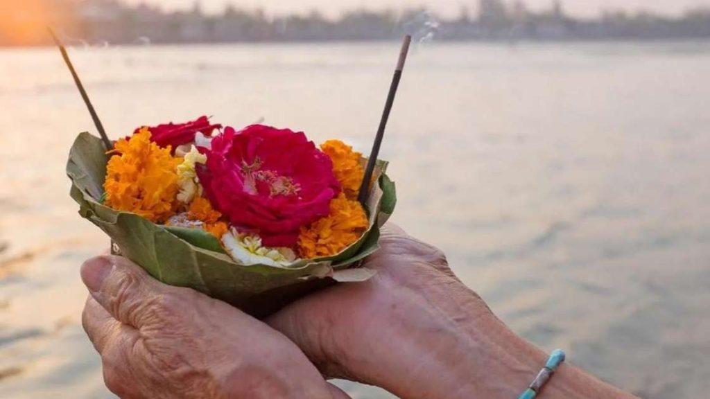 Hands gently holding a leaf bowl with a circular arrangement of red and orange flowers against the serene backdrop of a sunset over a river, symbolizing the tranquility and respect observed during Pitru Paksha.